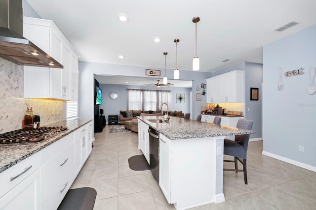 kitchen featuring light stone countertops, wall chimney exhaust hood, sink, a center island with sink, and white cabinets