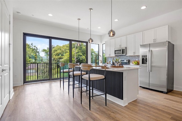 kitchen with stainless steel appliances, a kitchen island with sink, decorative light fixtures, white cabinets, and light hardwood / wood-style floors