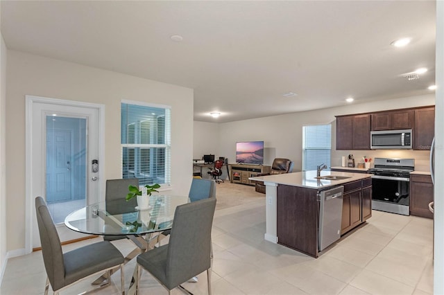 kitchen with a center island with sink, sink, light tile patterned floors, appliances with stainless steel finishes, and dark brown cabinetry