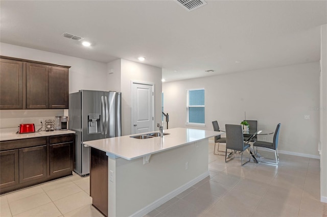 kitchen with stainless steel refrigerator with ice dispenser, dark brown cabinetry, a kitchen island with sink, sink, and light tile patterned floors