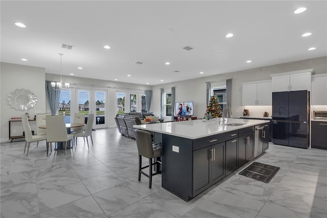 kitchen with a kitchen island with sink, black fridge, sink, decorative light fixtures, and white cabinetry