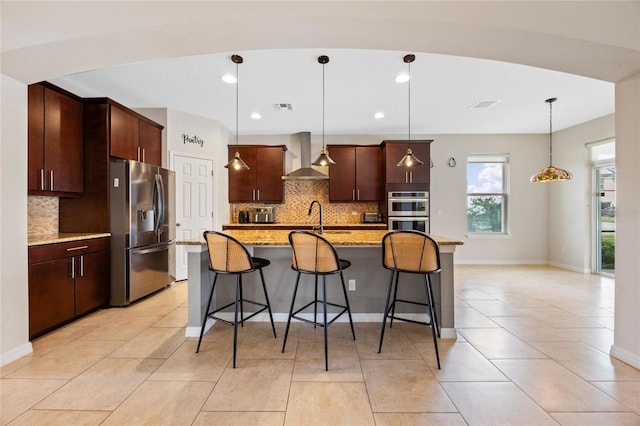 kitchen with wall chimney exhaust hood, backsplash, an island with sink, pendant lighting, and appliances with stainless steel finishes