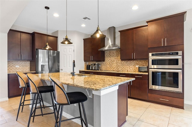 kitchen with sink, hanging light fixtures, stainless steel appliances, wall chimney range hood, and a kitchen island with sink