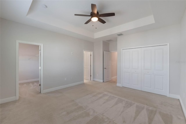 unfurnished bedroom with a walk in closet, ceiling fan, a tray ceiling, light colored carpet, and a closet