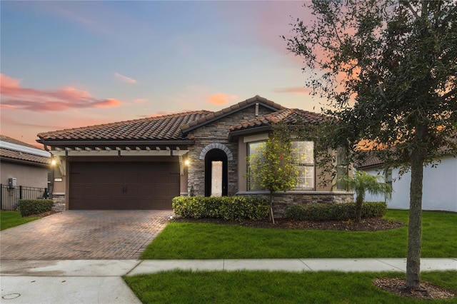 mediterranean / spanish-style house featuring decorative driveway, a tile roof, a lawn, a garage, and stone siding