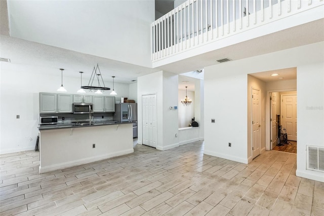 kitchen with gray cabinetry, a high ceiling, decorative light fixtures, a kitchen island with sink, and appliances with stainless steel finishes