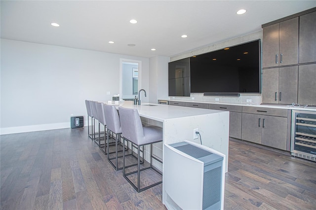 kitchen featuring beverage cooler, a breakfast bar, a kitchen island with sink, a sink, and dark wood-type flooring