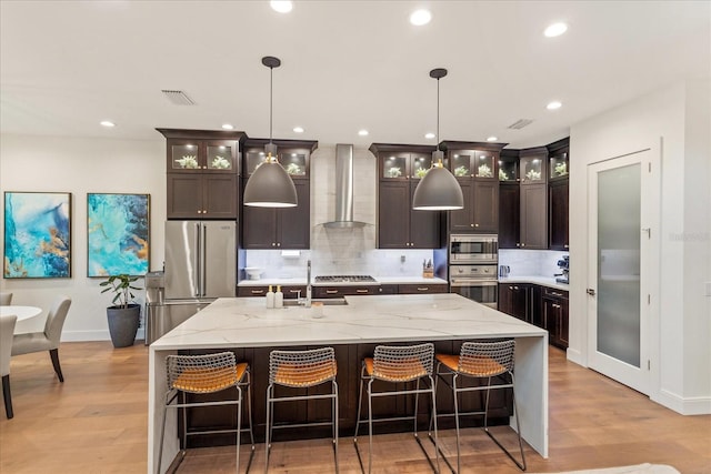 kitchen featuring a kitchen island with sink, hanging light fixtures, wall chimney exhaust hood, and stainless steel appliances