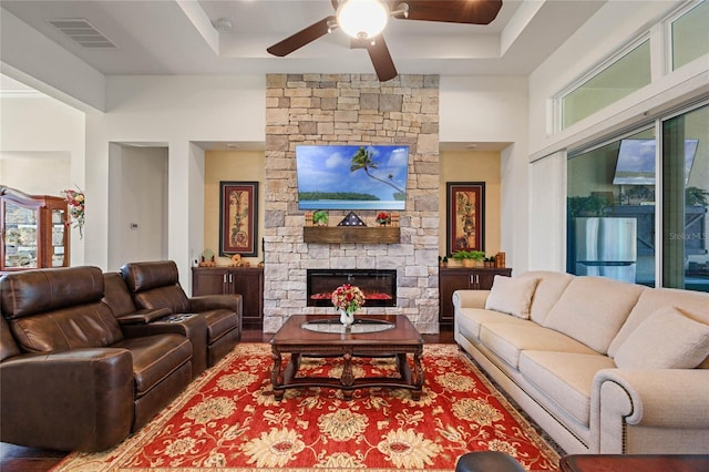 living area with a raised ceiling, visible vents, ceiling fan, a stone fireplace, and wood finished floors