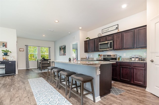 kitchen with hardwood / wood-style floors, a kitchen island with sink, appliances with stainless steel finishes, a kitchen bar, and dark brown cabinetry