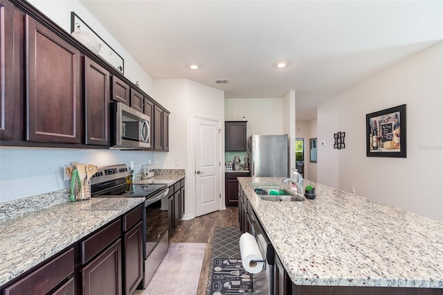 kitchen with light stone counters, hardwood / wood-style floors, an island with sink, dark brown cabinets, and appliances with stainless steel finishes