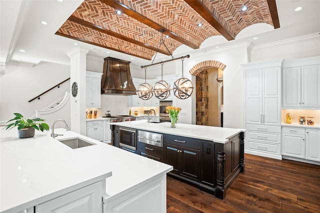 kitchen featuring brick ceiling, a large island with sink, white cabinets, and premium range hood