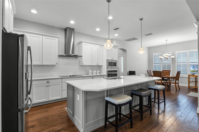 kitchen with wall chimney exhaust hood, stainless steel appliances, a kitchen island with sink, white cabinetry, and hanging light fixtures