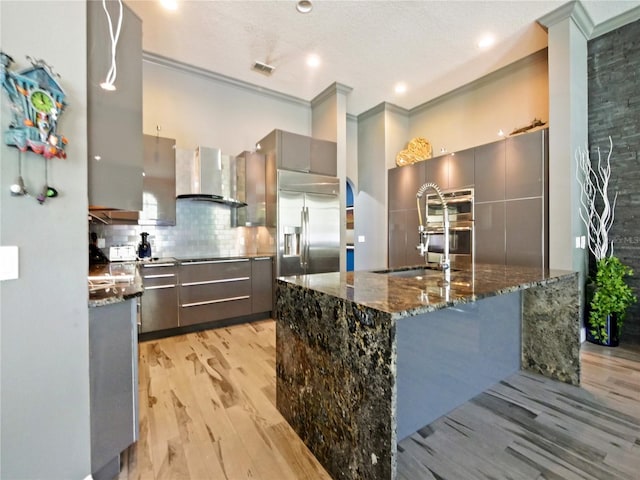 kitchen with stainless steel built in refrigerator, tasteful backsplash, light wood-type flooring, dark stone countertops, and wall chimney range hood