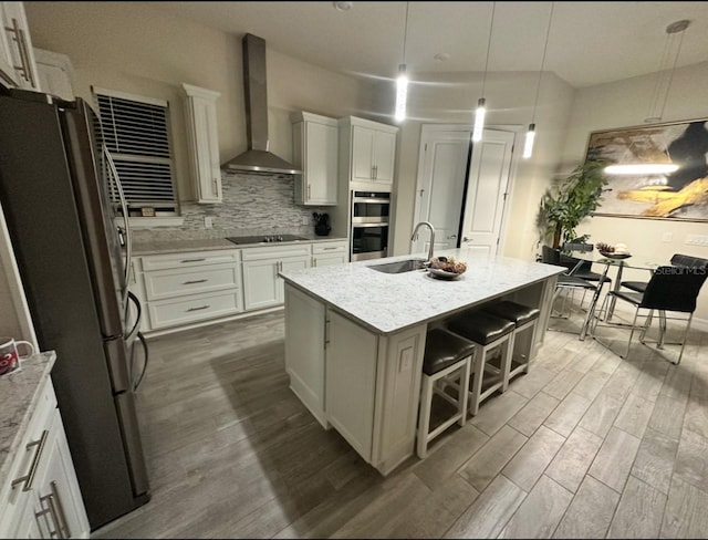 kitchen with stainless steel appliances, a kitchen island with sink, sink, wall chimney range hood, and pendant lighting