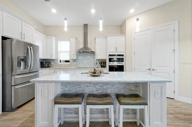 kitchen featuring a center island with sink, white cabinets, appliances with stainless steel finishes, wall chimney range hood, and tasteful backsplash