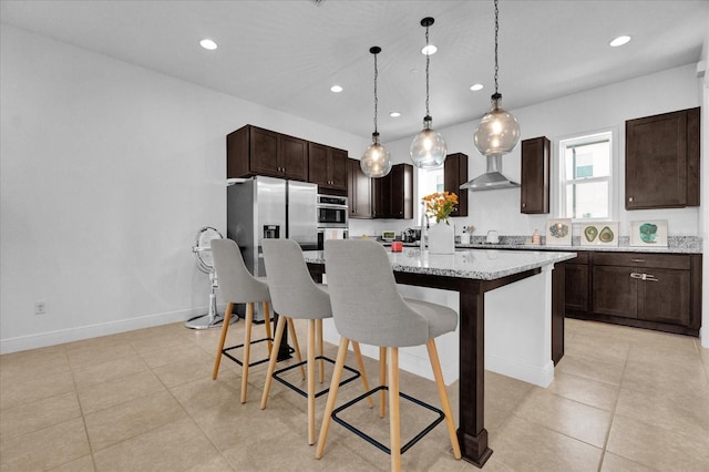 kitchen featuring light stone countertops, wall chimney range hood, a kitchen breakfast bar, an island with sink, and decorative light fixtures