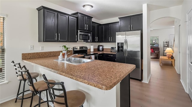 kitchen featuring arched walkways, dark cabinets, stainless steel appliances, a peninsula, and a sink