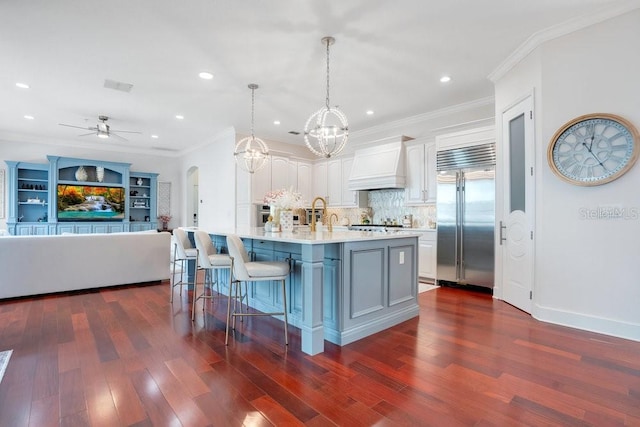 kitchen featuring hanging light fixtures, built in refrigerator, an island with sink, white cabinets, and custom exhaust hood