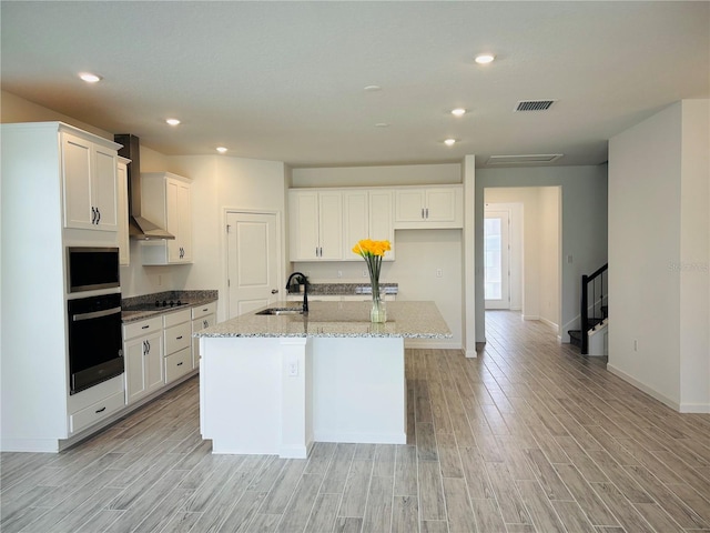 kitchen featuring sink, oven, gas stovetop, a center island with sink, and wall chimney range hood