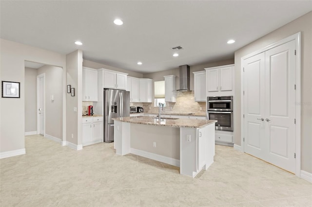 kitchen featuring appliances with stainless steel finishes, light stone counters, wall chimney exhaust hood, a kitchen island with sink, and white cabinetry