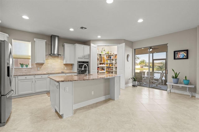 kitchen with wall chimney exhaust hood, light stone counters, a center island with sink, white cabinets, and appliances with stainless steel finishes