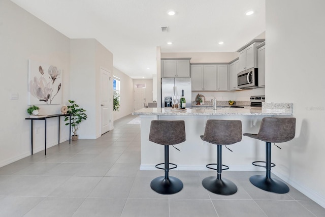 kitchen with light stone countertops, stainless steel appliances, kitchen peninsula, a kitchen bar, and gray cabinets