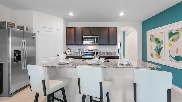 kitchen featuring a breakfast bar, a center island with sink, light stone countertops, dark brown cabinetry, and stainless steel appliances