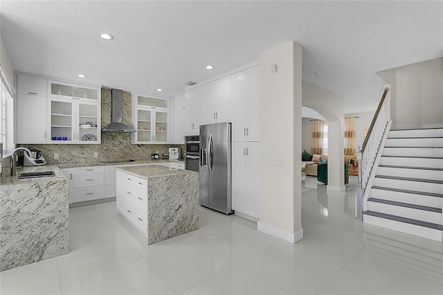 kitchen with wall chimney range hood, sink, light stone countertops, a kitchen island, and stainless steel fridge with ice dispenser