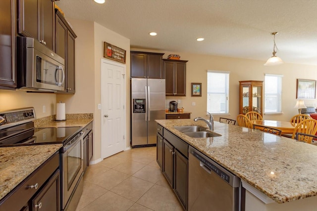 kitchen with light stone countertops, sink, hanging light fixtures, stainless steel appliances, and a kitchen island with sink