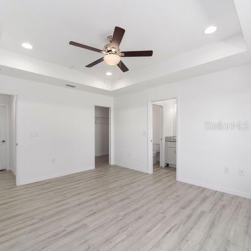 unfurnished bedroom with ceiling fan, a raised ceiling, light wood-type flooring, and ensuite bathroom