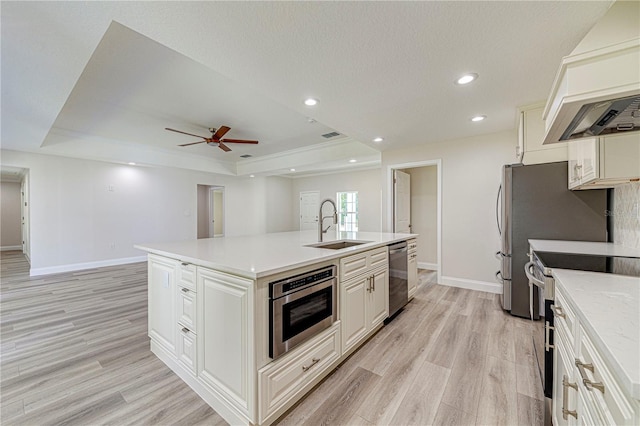 kitchen with a tray ceiling, stainless steel appliances, a center island with sink, and light hardwood / wood-style floors