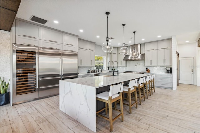 kitchen featuring light stone countertops, pendant lighting, a large island with sink, wall chimney range hood, and stainless steel built in refrigerator