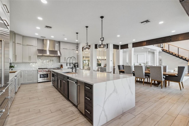 kitchen featuring a large island, stainless steel appliances, hanging light fixtures, light stone counters, and wall chimney range hood