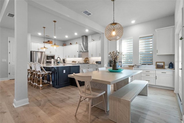 kitchen featuring a kitchen breakfast bar, wall chimney exhaust hood, a kitchen island with sink, white cabinets, and hanging light fixtures