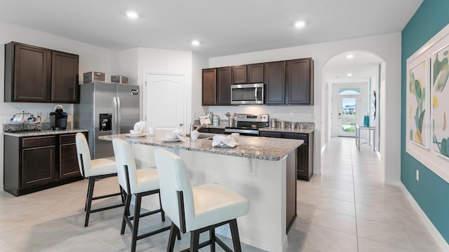 kitchen featuring a kitchen bar, light stone counters, dark brown cabinets, stainless steel appliances, and a kitchen island with sink