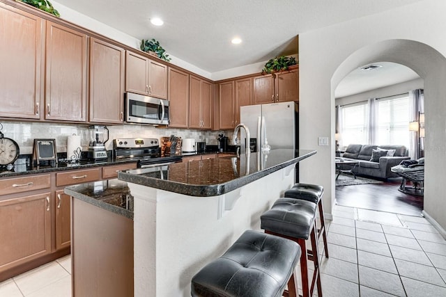 kitchen with a center island with sink, a kitchen breakfast bar, dark stone countertops, light tile patterned flooring, and stainless steel appliances