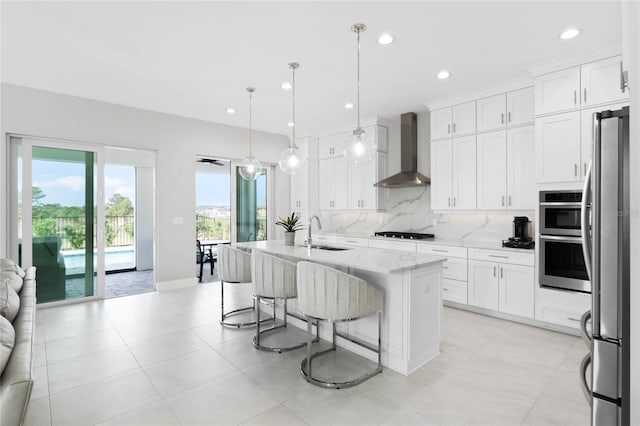 kitchen featuring a center island with sink, sink, hanging light fixtures, wall chimney exhaust hood, and white cabinetry