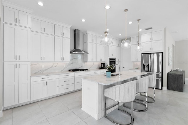 kitchen with a center island with sink, hanging light fixtures, wall chimney exhaust hood, stainless steel fridge, and white cabinetry