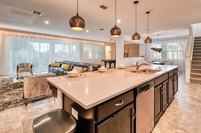 kitchen with light stone countertops, dishwasher, sink, hanging light fixtures, and a center island with sink