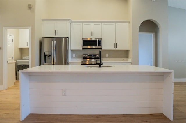 kitchen featuring white cabinets, a towering ceiling, an island with sink, stainless steel appliances, and washer / clothes dryer