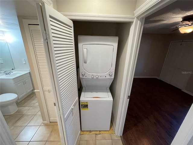 clothes washing area featuring light tile patterned floors, ceiling fan, laundry area, stacked washer / dryer, and a sink