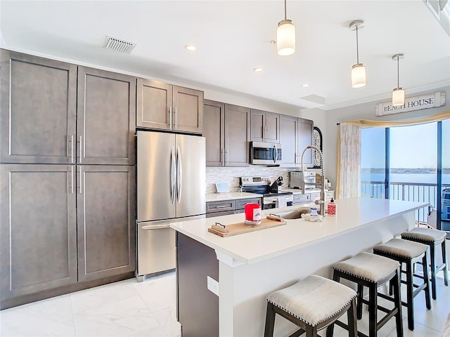 kitchen with stainless steel appliances, an island with sink, decorative backsplash, hanging light fixtures, and a water view