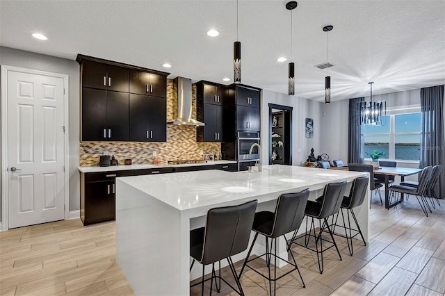 kitchen featuring a kitchen breakfast bar, pendant lighting, decorative backsplash, wall chimney range hood, and a kitchen island with sink