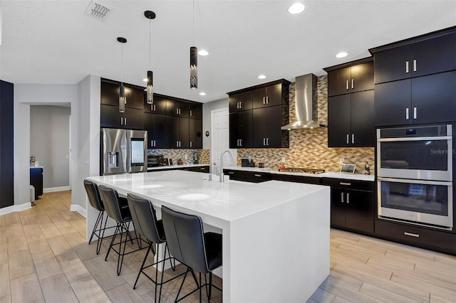 kitchen featuring a center island with sink, appliances with stainless steel finishes, hanging light fixtures, wall chimney exhaust hood, and sink