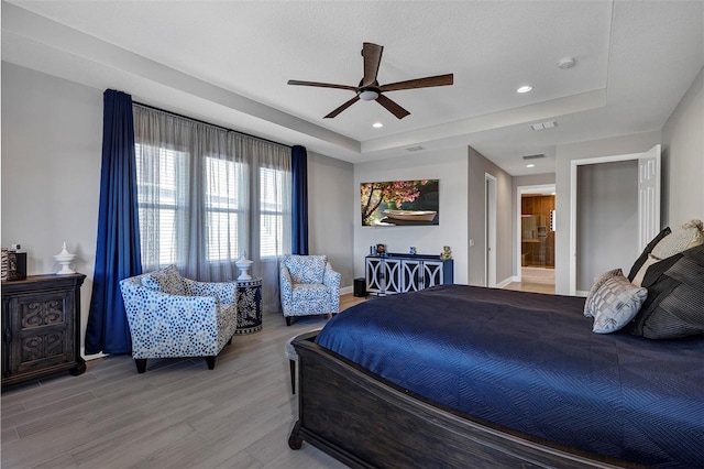 bedroom with ensuite bathroom, a raised ceiling, ceiling fan, and light wood-type flooring