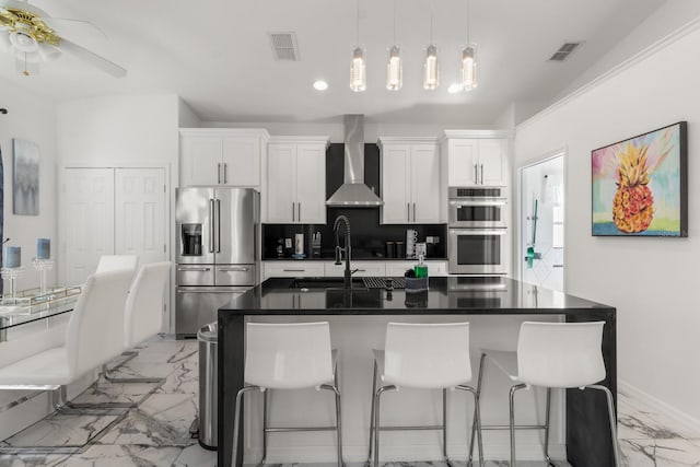 kitchen with stainless steel appliances, wall chimney range hood, a kitchen breakfast bar, an island with sink, and white cabinets