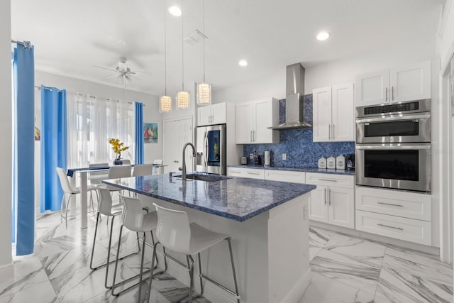 kitchen featuring ceiling fan, sink, stainless steel appliances, wall chimney range hood, and a center island with sink