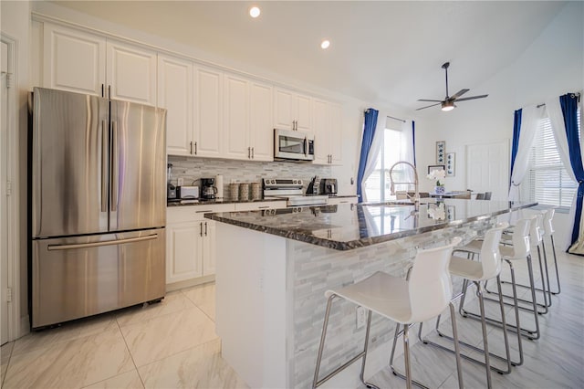 kitchen featuring ceiling fan, dark stone countertops, a kitchen island with sink, white cabinets, and appliances with stainless steel finishes