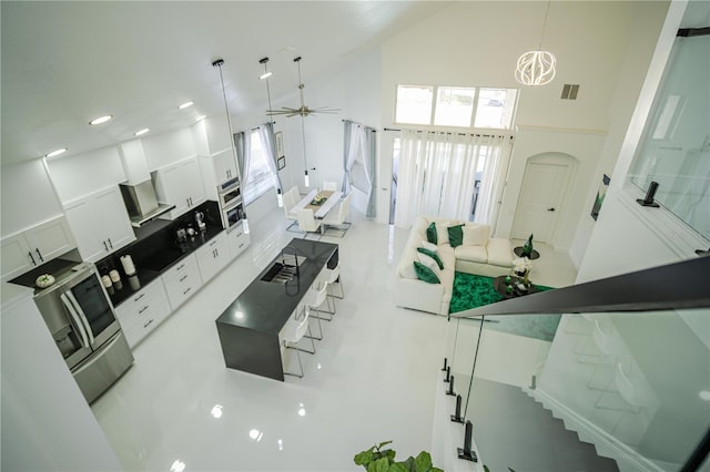 kitchen with a towering ceiling, ceiling fan with notable chandelier, wall chimney range hood, decorative light fixtures, and white cabinetry